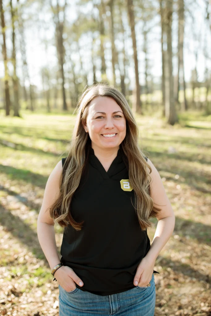 Woman in a black sleeveless top standing outdoors in a wooded area, smiling warmly at the camera.