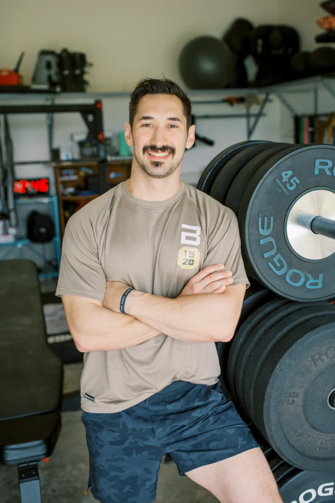 Man in a tan workout shirt standing in a gym, leaning against a stack of weight plates with arms crossed.