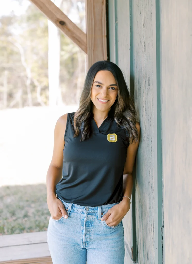 Woman in a black sleeveless top and jeans leaning casually against a wooden wall, smiling.