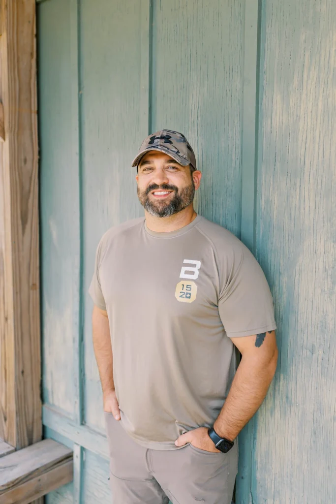 Smiling man in a tan t-shirt and camo cap leaning against a rustic wooden wall.