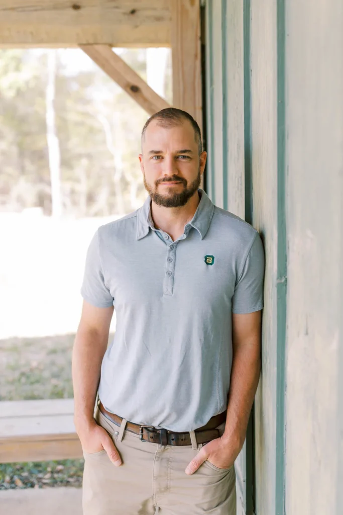 Man in a light gray polo shirt leaning against a wooden wall outdoors, looking confidently at the camera.