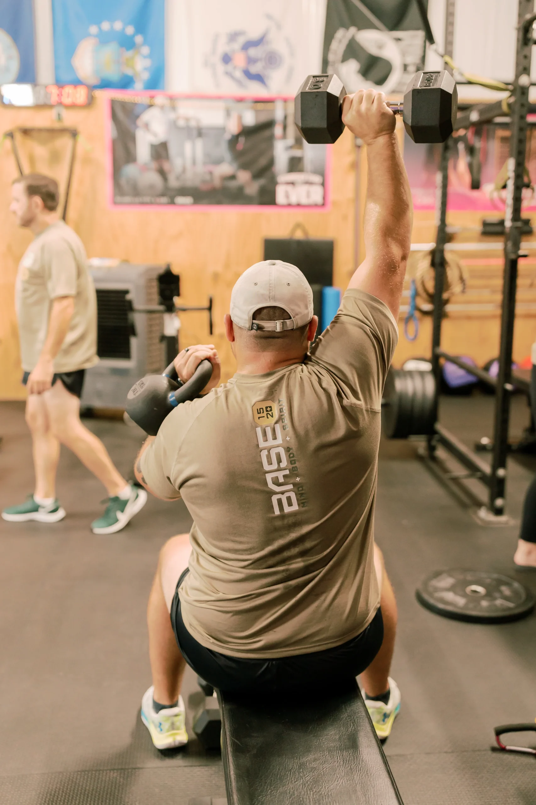 Man seated on a workout bench, performing a one-arm overhead dumbbell press in a gym setting.