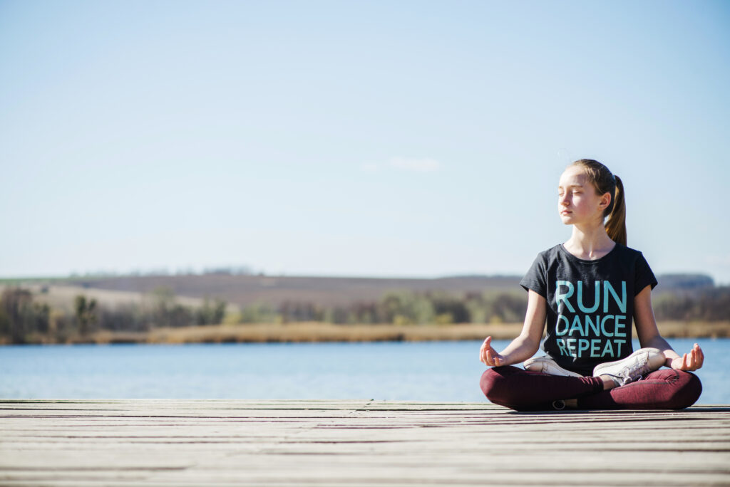 Young girl meditating in a cross-legged position on a wooden dock by a lake, with eyes closed.