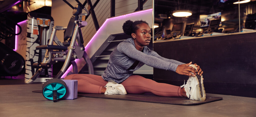 Woman in a gym sitting on a mat, stretching forward to touch her feet with workout equipment nearby.