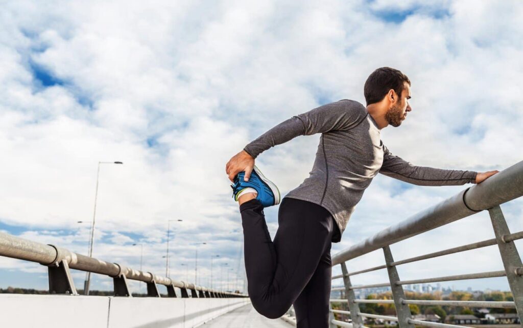 Man stretching his quadriceps on a bridge railing while wearing athletic wear.