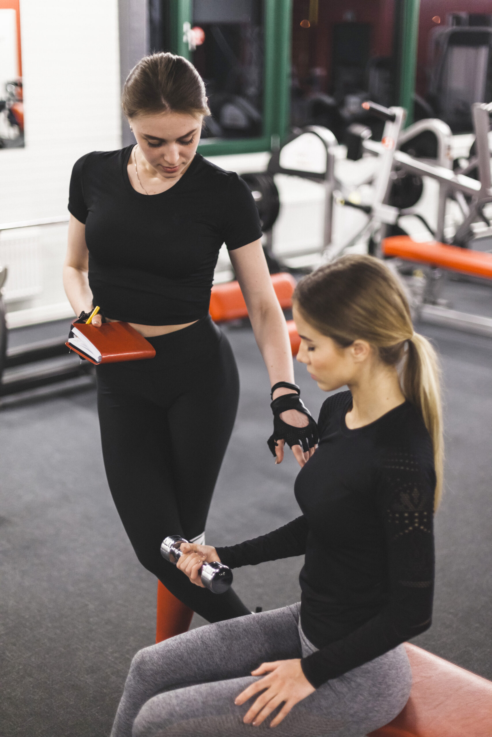 Female trainer supervising another woman lifting a dumbbell while taking notes in a gym.