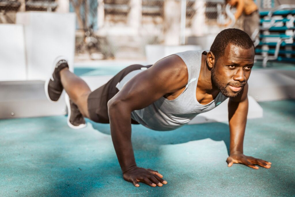 Man in a gray tank top performing push-ups outdoors on a turquoise workout surface.