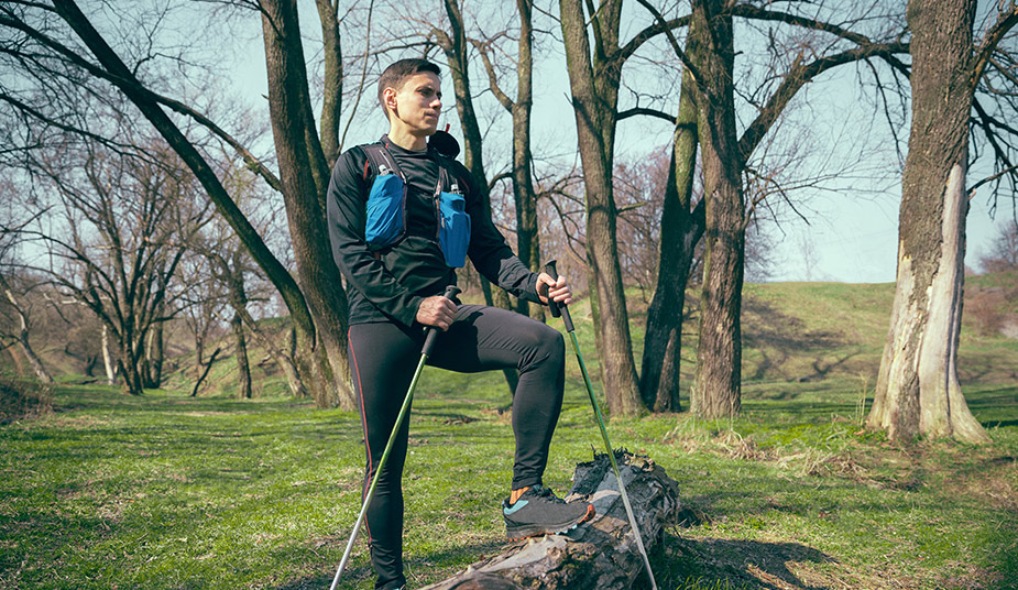 Person wearing a black outfit with a hydration pack, standing in a wooded area holding trekking poles.