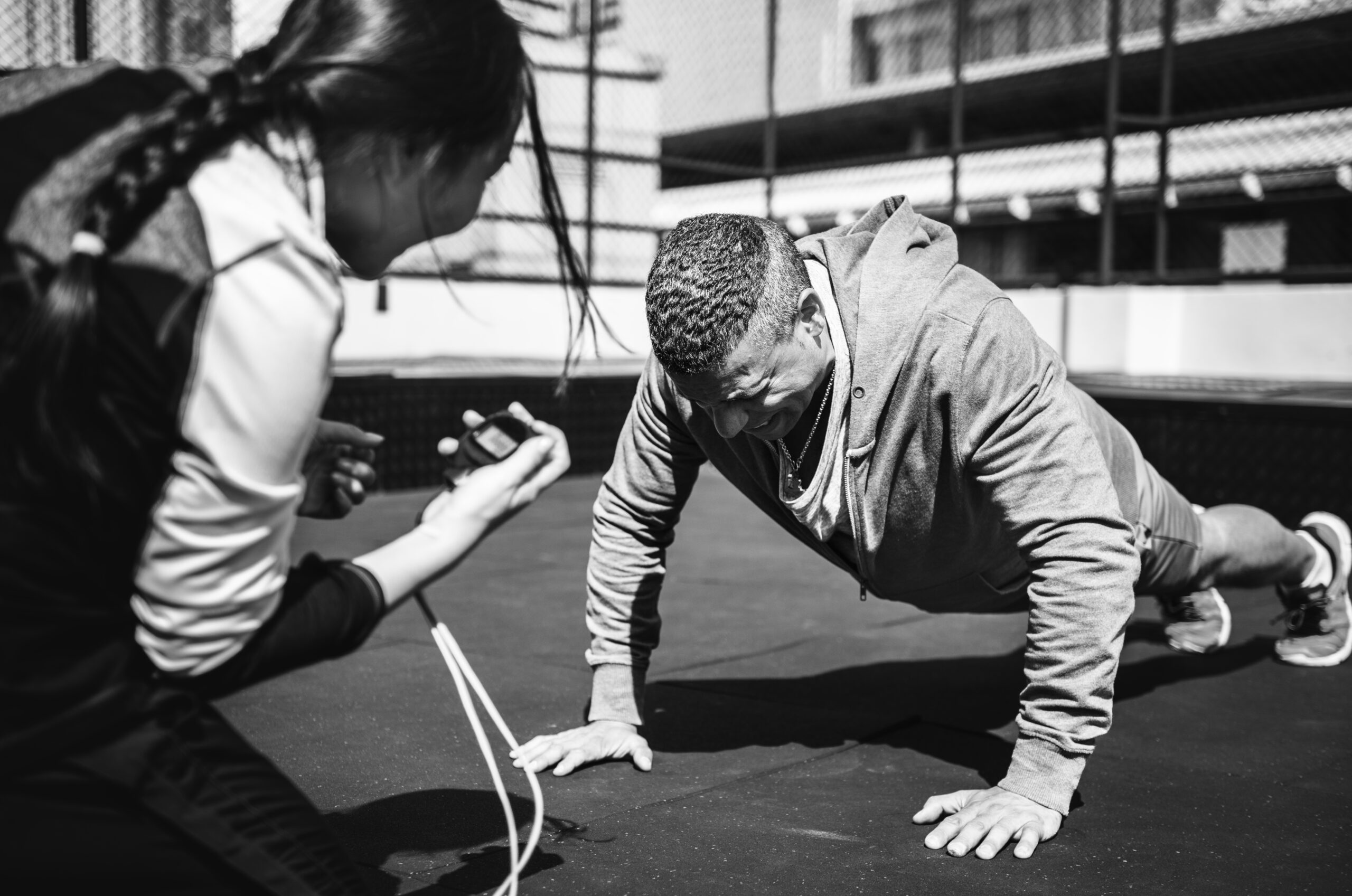 Black and white image of a man doing push-ups outdoors while a trainer observes and times him.